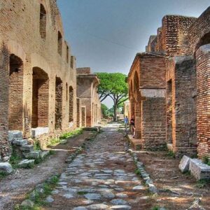 L'immagine mostra una strada acciottolata fiancheggiata da antichi edifici in mattoni rossi, identificati come le caratteristiche insulae (caseggiati a più piani) nel Parco Archeologico di Ostia Antica.