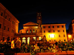 Vista notturna sulla piazza di Santa Maria in Trastevere animata dalla movida locale alla quale si aggiungo turisti da tutto il mondo, un ritrovo affascinante e multiculturale.