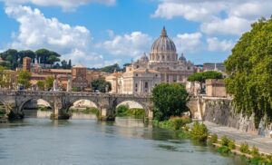 The image captures a sweeping panoramic view of Rome, with the Tiber River, a historic bridge, and the majestic dome of St. Peter's Basilica rising in the distance. The Tiber, the river that winds through the Eternal City, is visible in the foreground with its distinctive embankments and riverfront quays. The historic bridges spanning the Tiber are masonry arch structures linking the two shores—a hallmark of Roman engineering and architecture. Overlooking it all is Michelangelo's iconic dome of St. Peter's, a landmark that dominates the Vatican City skyline and defines Rome's horizon.