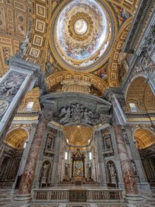 The image depicts the interior of St. Peter's Basilica in the Vatican, offering a view from below of one of the subsidiary arches and the dome, which is decorated with mosaics. The massive marble pillars feature columns with Corinthian capitals, and are surmounted by white marble angel sculptures and ornamental stuccoes that frame the vaulted ceilings.