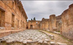 L'immagine mostra uno scorcio del parco archeologico di Ostia Antica, l'antico porto commerciale di Roma, con la tipica pavimentazione in pietra e gli edifici in mattoni rossi risalenti all'epoca imperiale.